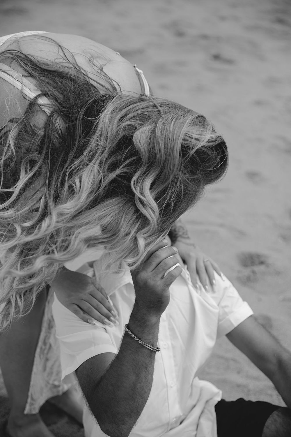 Black and white photo of a couple embracing on a sandy beach, woman with curly blonde hair leaning in for a kiss.