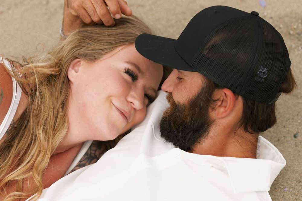 A smiling couple lying together on a beach, gazing at each other, woman with blonde hair and man with beard and cap.