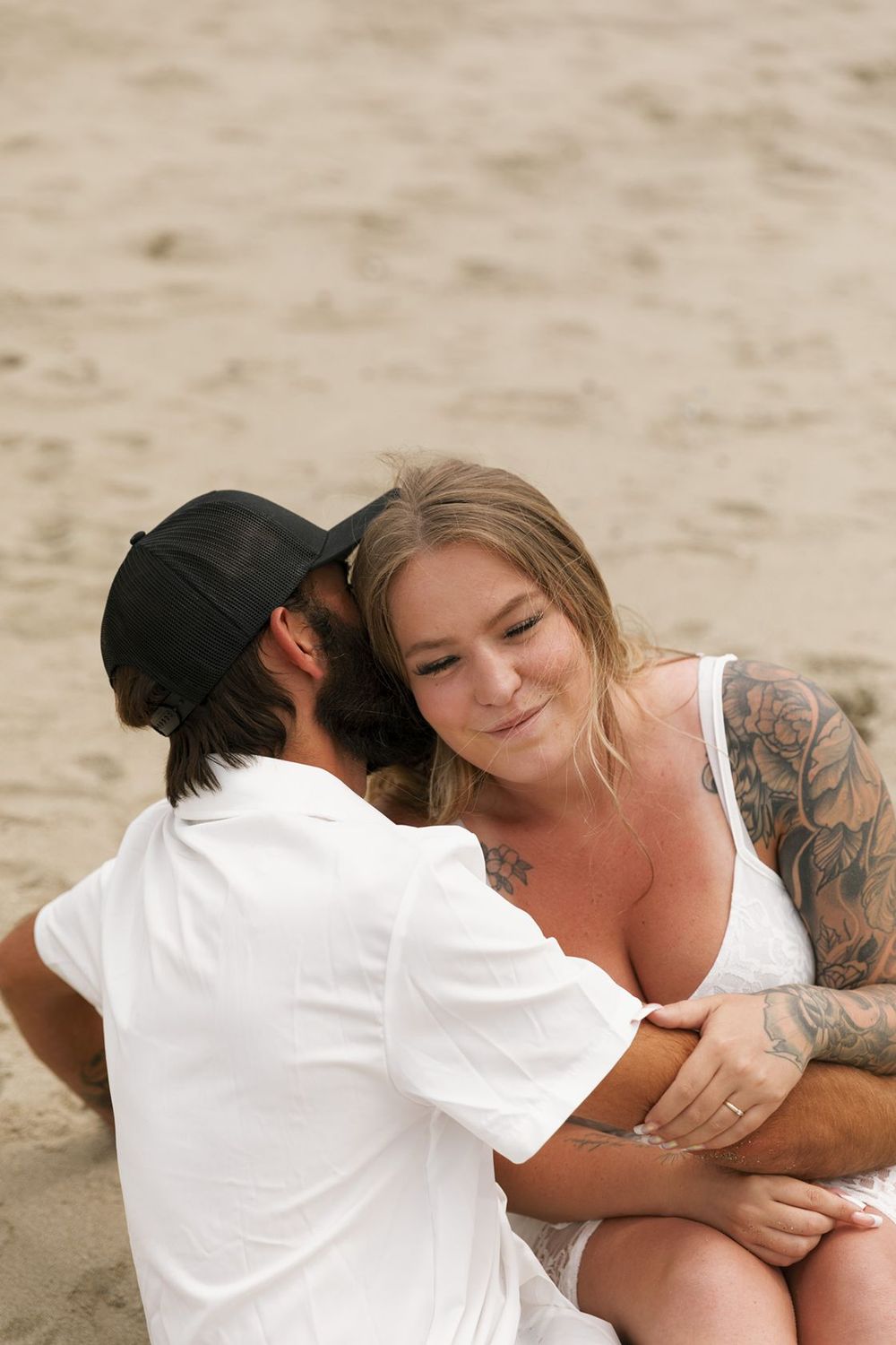 Couple sharing a tender moment on a sandy beach, woman with tattoos smiling warmly.