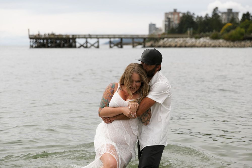 Romantic couple standing in shallow ocean water, woman in white dress embraced by tattooed partner near pier.