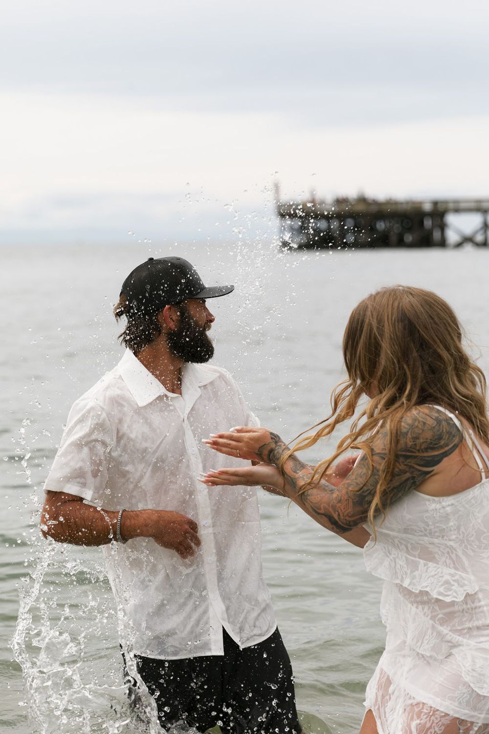 Couple in white outfits splashing water at the ocean shore, woman with tattoos and man with beard and cap.