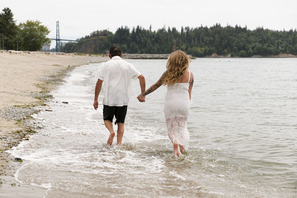 Couple holding hands walking along sandy shoreline with trees and bridge in background.