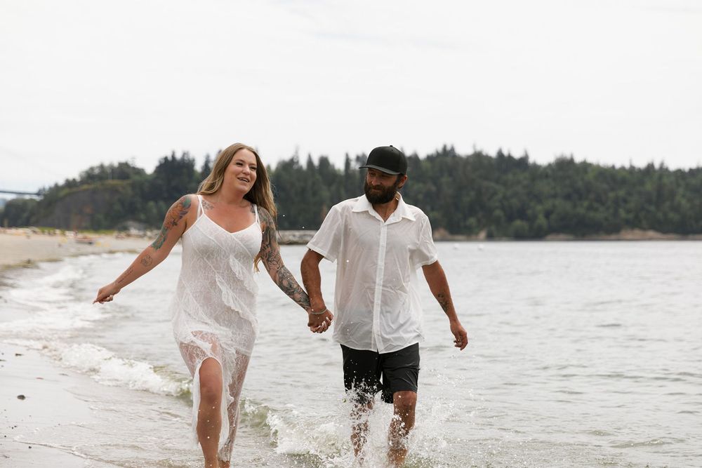 Couple holding hands and walking through ocean waves on a beach in white outfits.