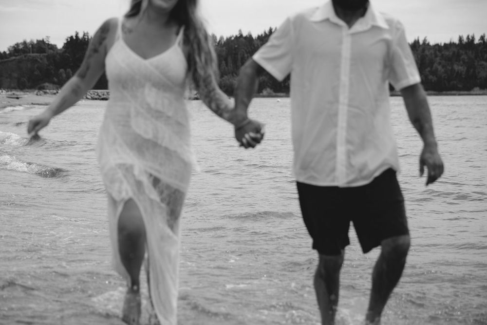 Couple in white outfits holding hands running along a beach shore in black and white photo.