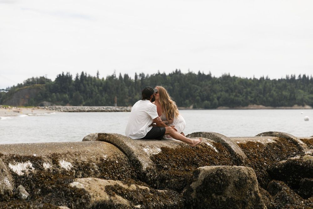 Couple sitting together on rocky shoreline overlooking scenic forested coastline.