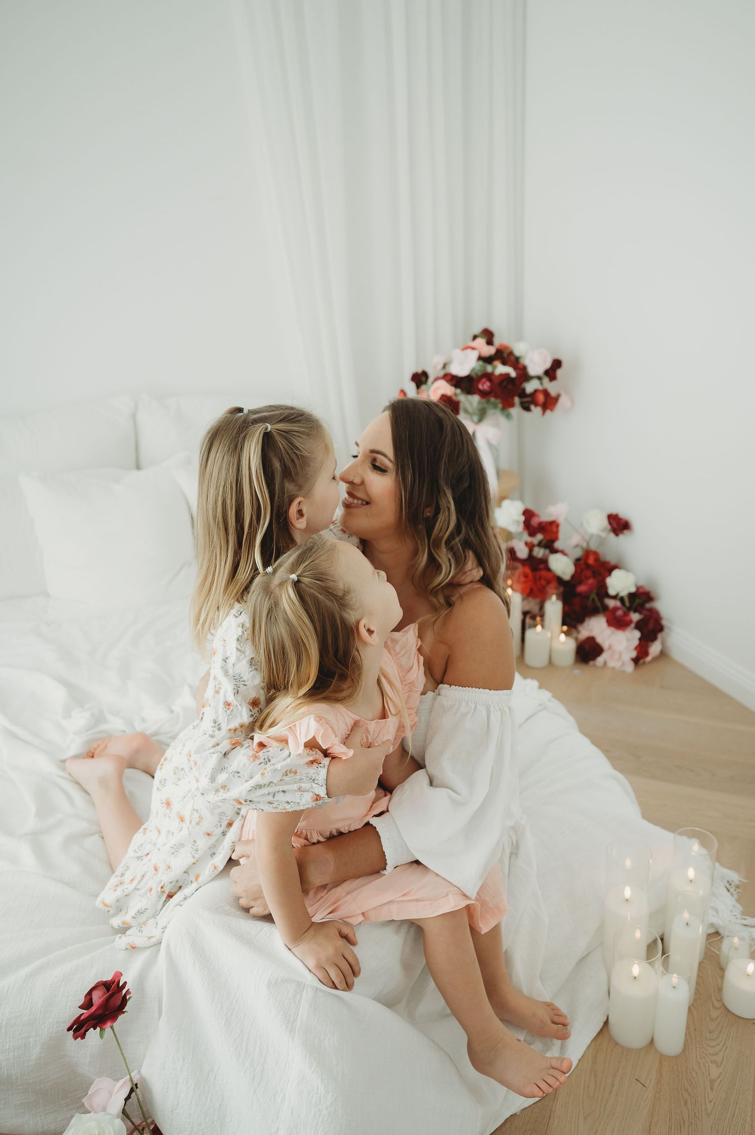 A mother sits on a white bed kissing her two young daughters surrounded by candles and red floral arrangements.