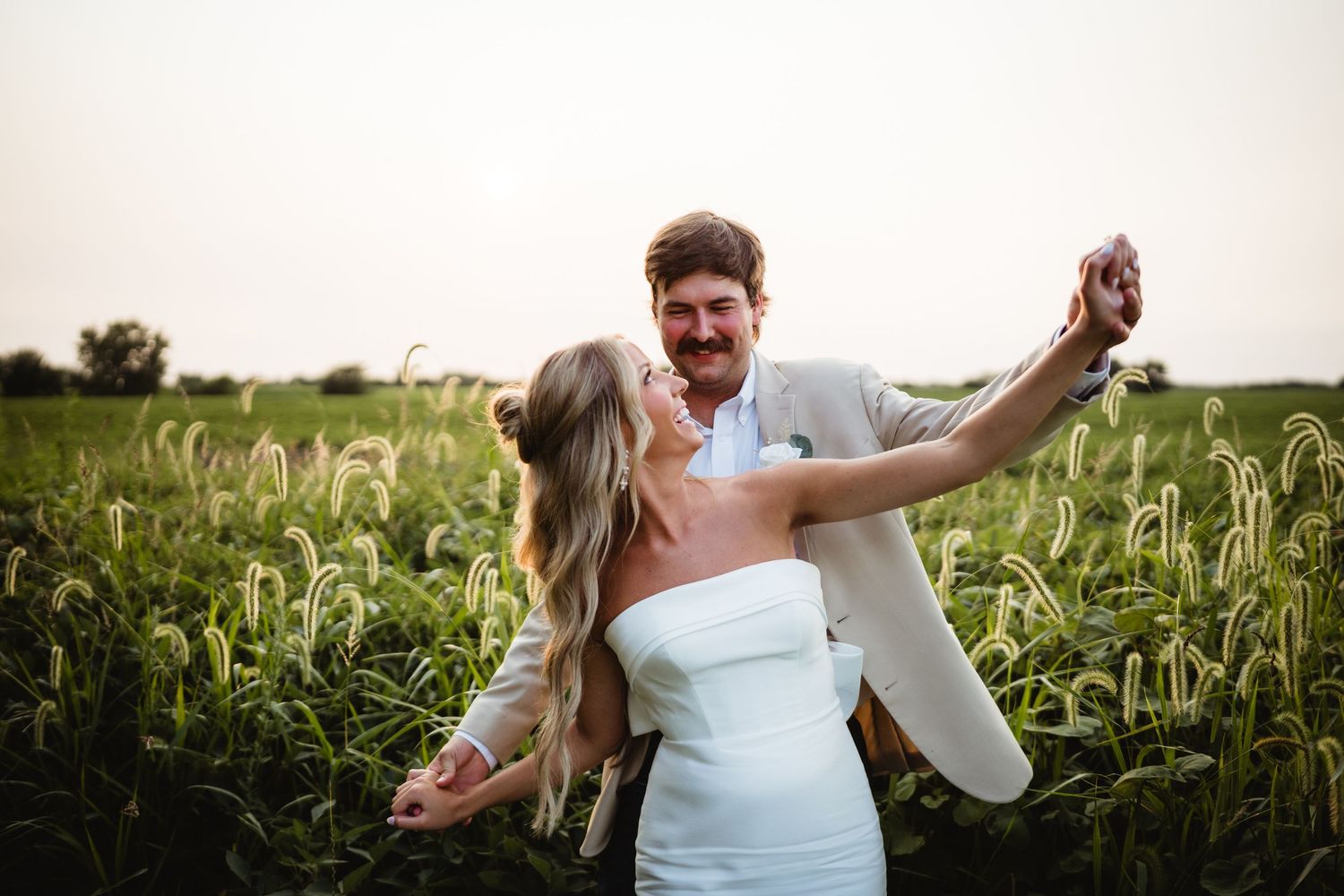 A joyful couple dances in a lush green field at sunset, bride in white dress, groom in tan suit.