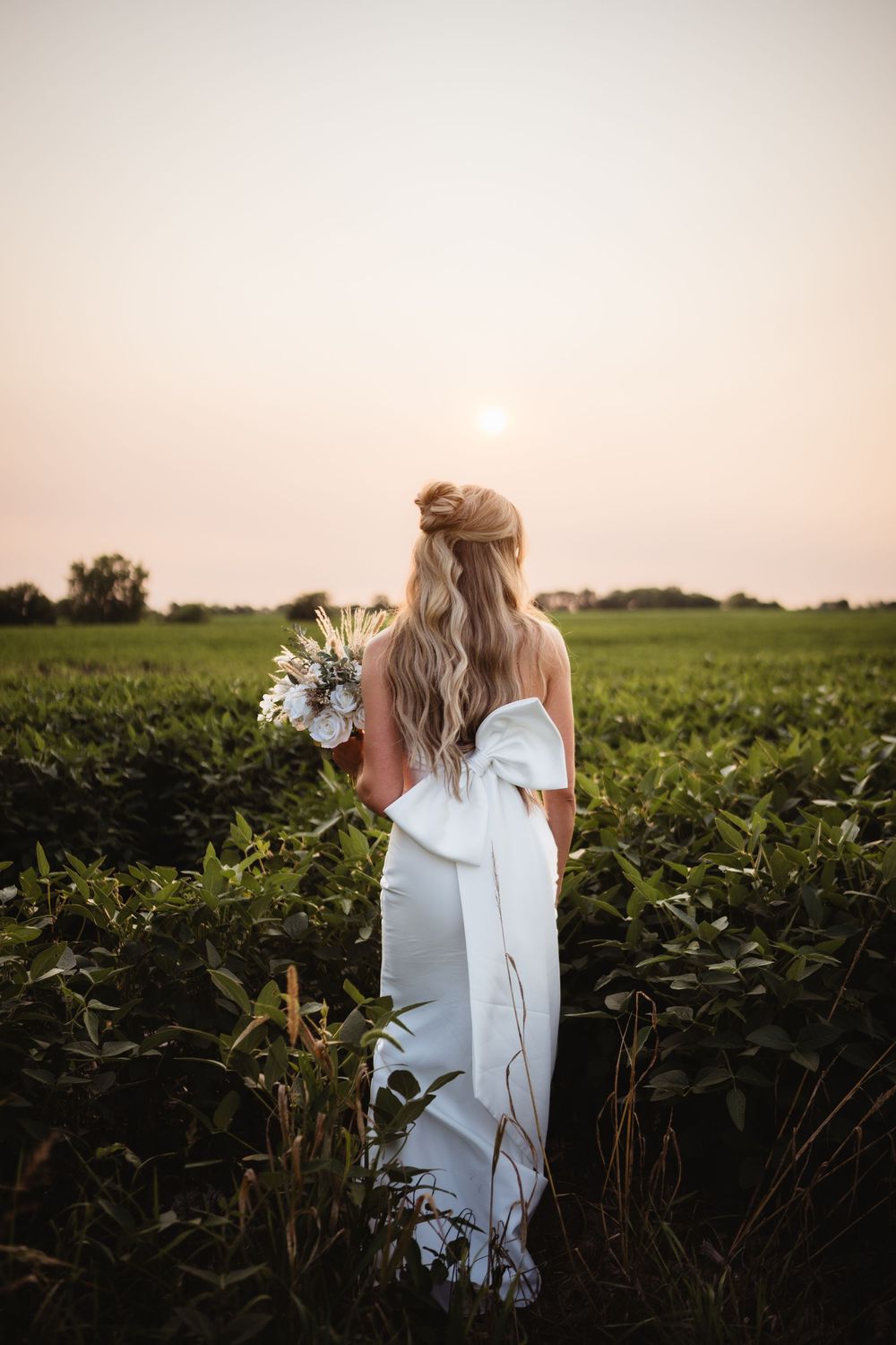 Bride in white off-shoulder gown with large bow holds bouquet in lush green field at golden sunset.