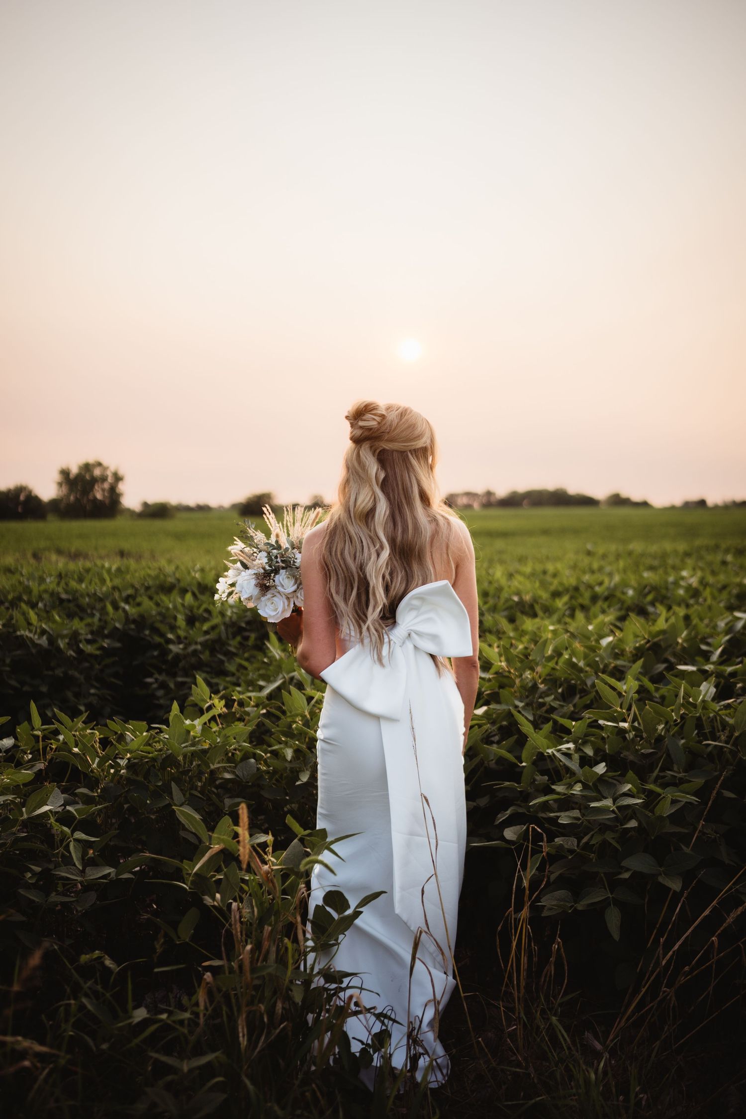 Bride in white off-shoulder gown with large bow holds bouquet in lush green field at golden sunset.
