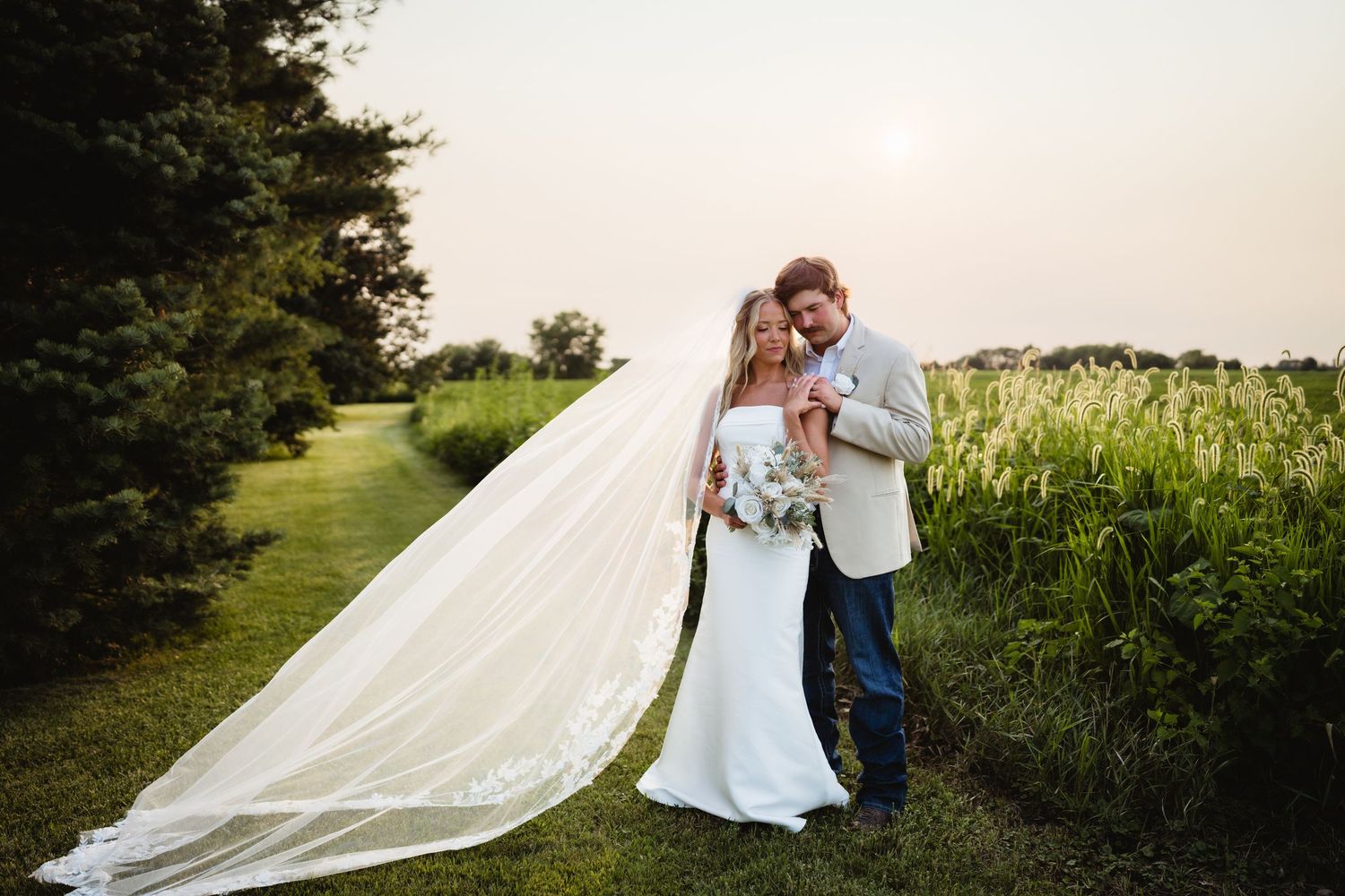 Bride and groom embrace at sunset in a field, her long flowing veil trailing behind them near lush greenery.
