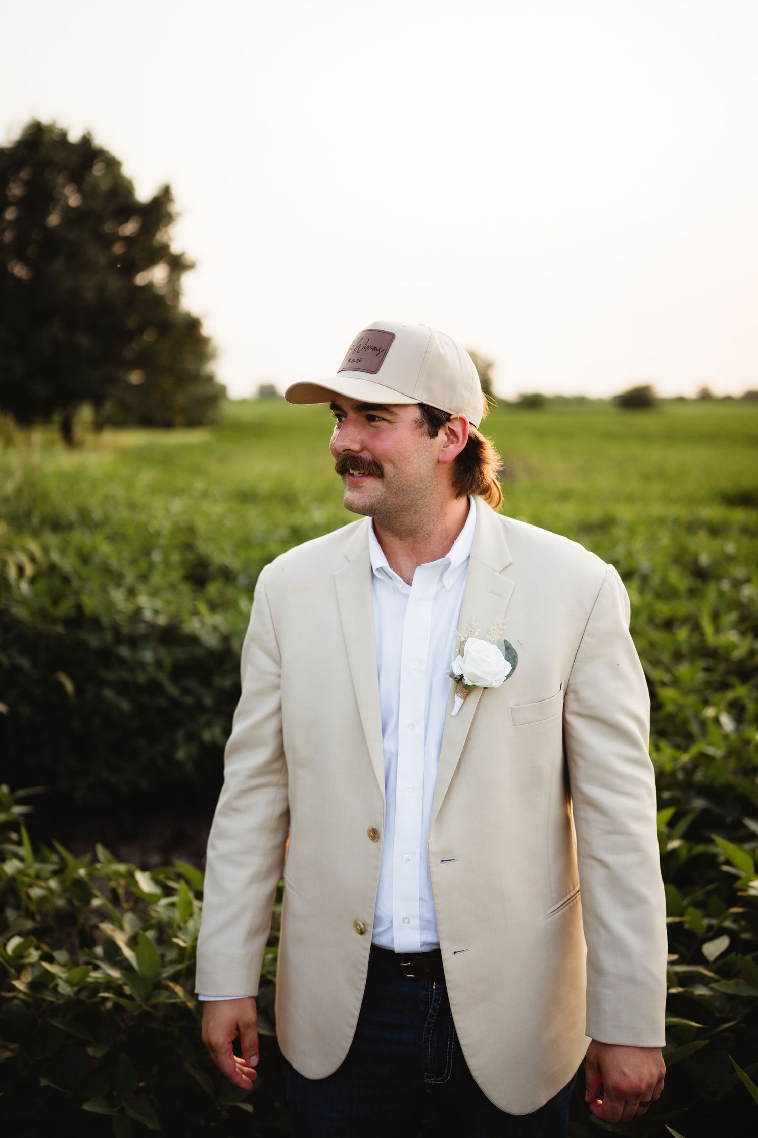 Groom in tan blazer and trucker cap with white boutonniere standing in green field at golden hour.