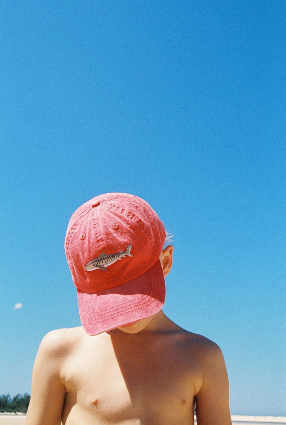 Shirtless boy at beach wearing a pink shark baseball cap against a clear blue sky.