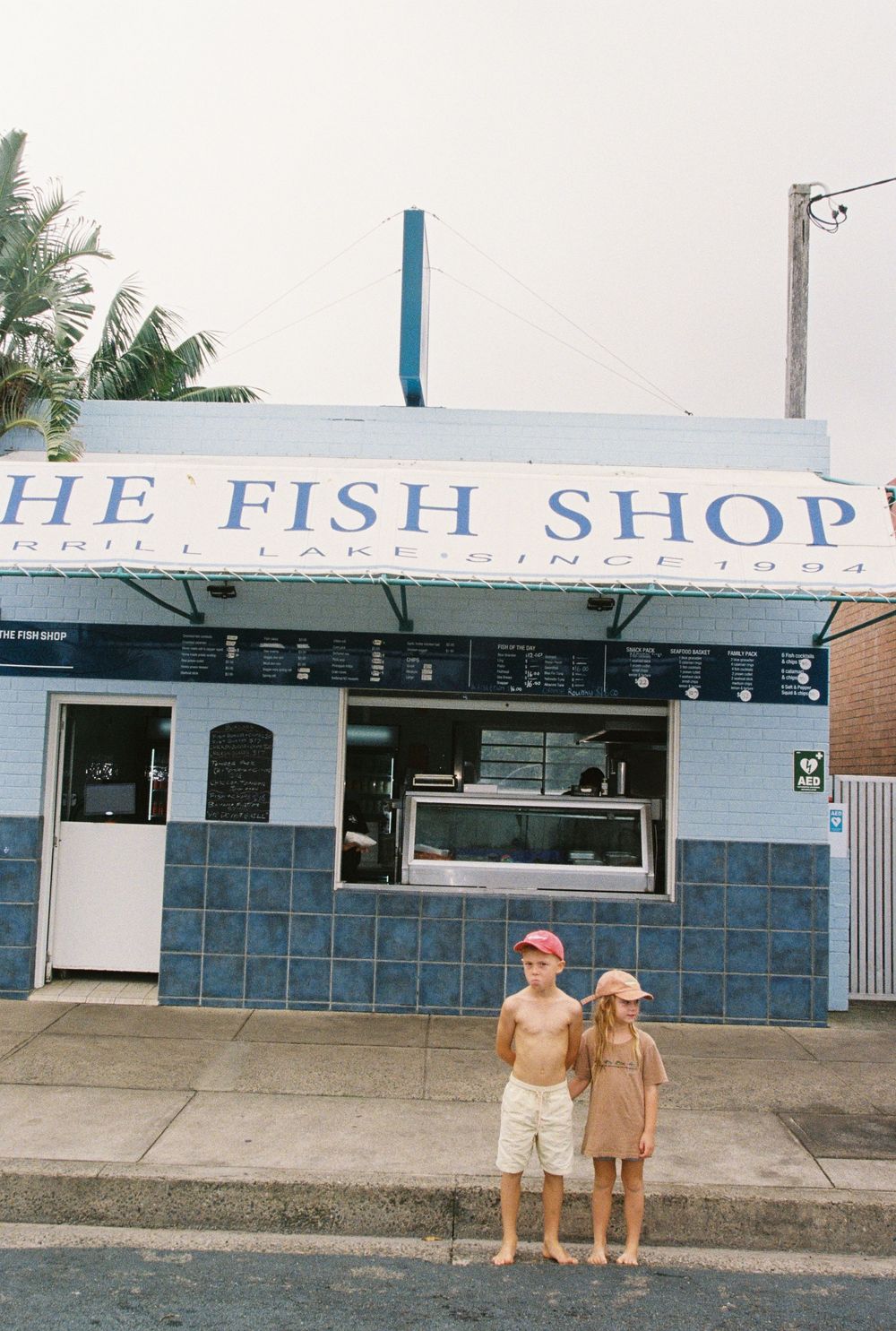 Two children stand in front of The Fish Shop, a blue-tiled seafood restaurant open since 1994, on a sunny day.