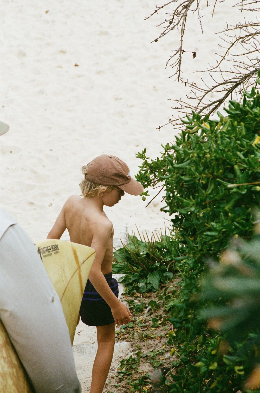 Young shirtless surfer boy carrying a yellow surfboard near beach vegetation wearing a pink cap.
