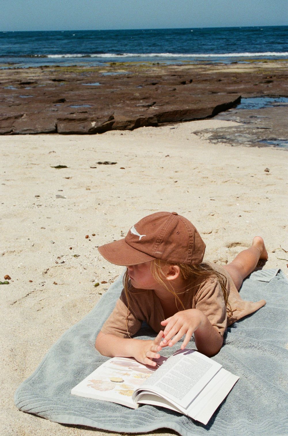 A young girl in a brown cap reads a book while lying on a towel on a sunny sandy beach.