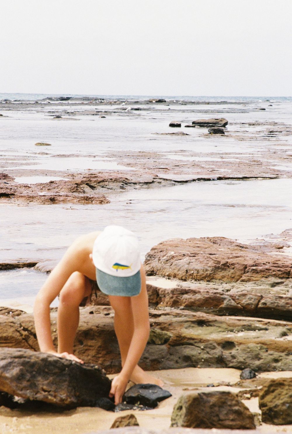 A young child in a white cap explores rocks and tide pools along a scenic rocky beach shoreline.