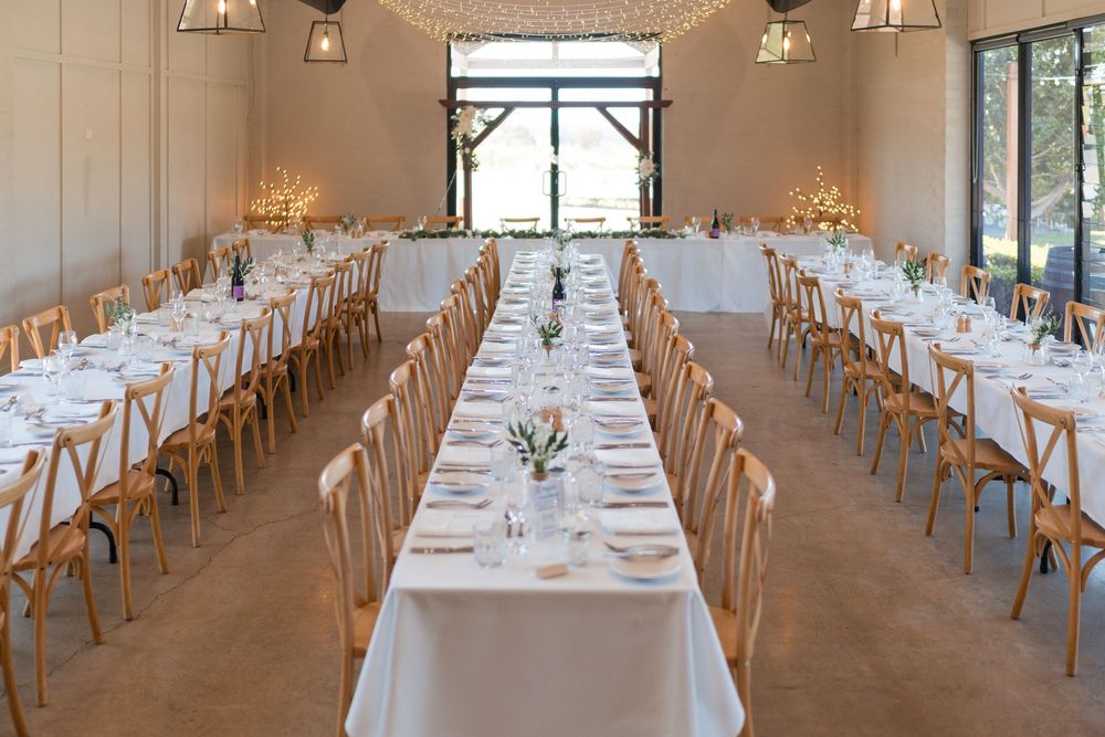 Elegant barn wedding reception with long white-clothed tables, cross-back chairs, and soft lighting.