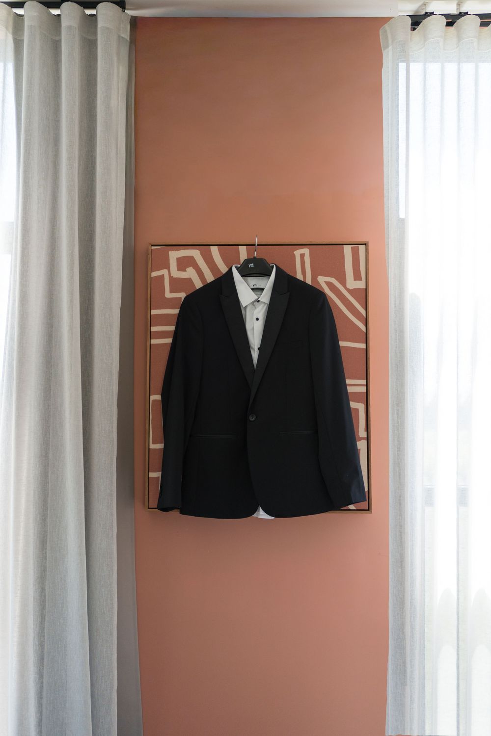 Black tuxedo jacket with white dress shirt on hanger displayed against terracotta wall with decorative artwork.