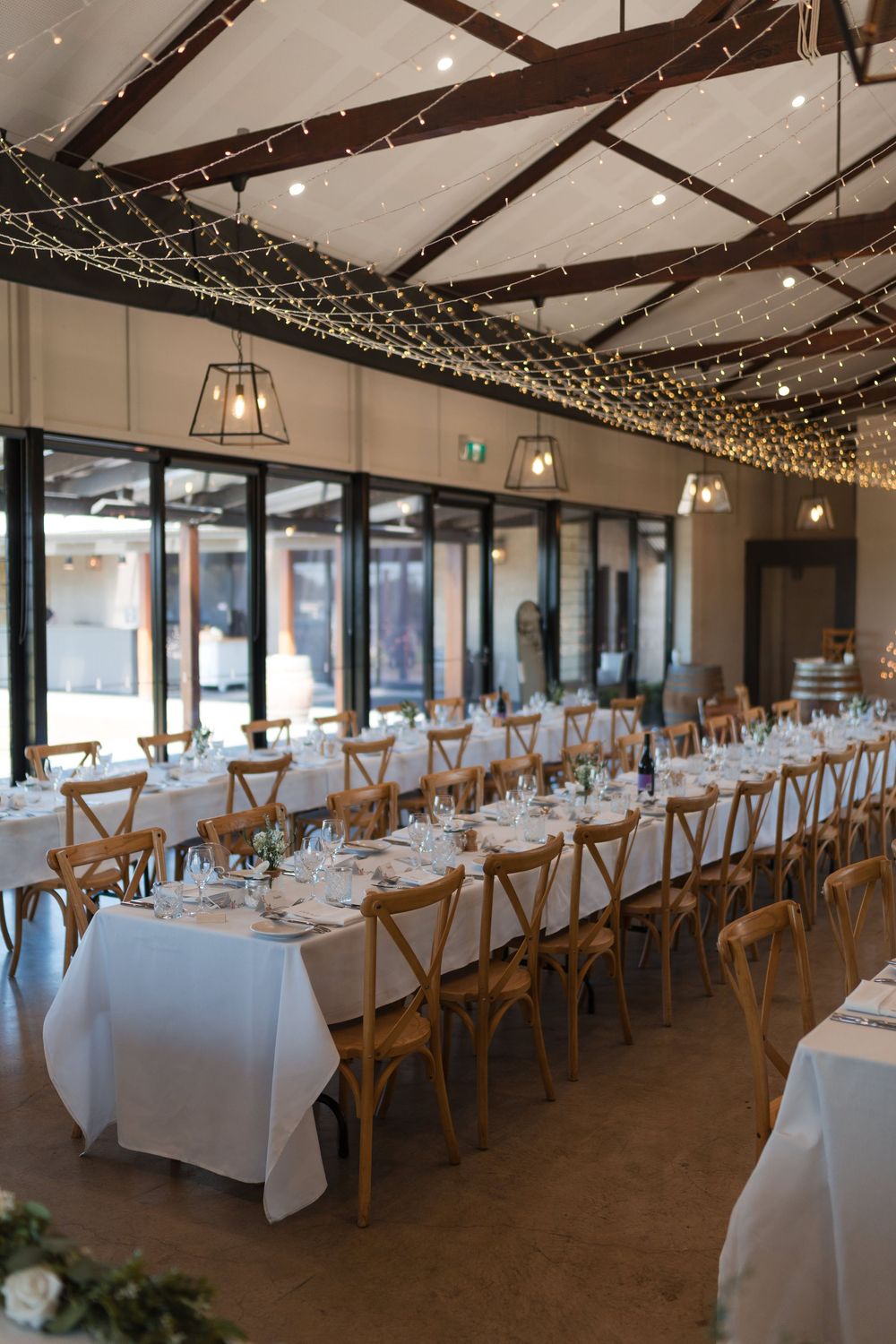 Elegant winery wedding reception hall with fairy lights, long white tables, wooden cross-back chairs, and pendant lighting.