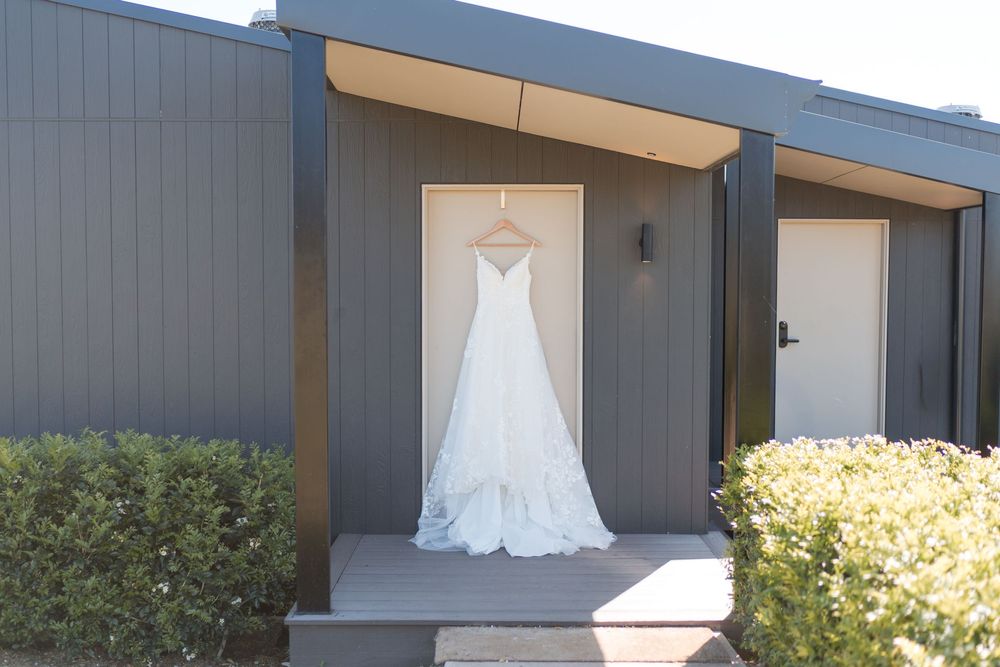 White lace wedding dress hanging on a door of a modern grey building surrounded by green shrubs.