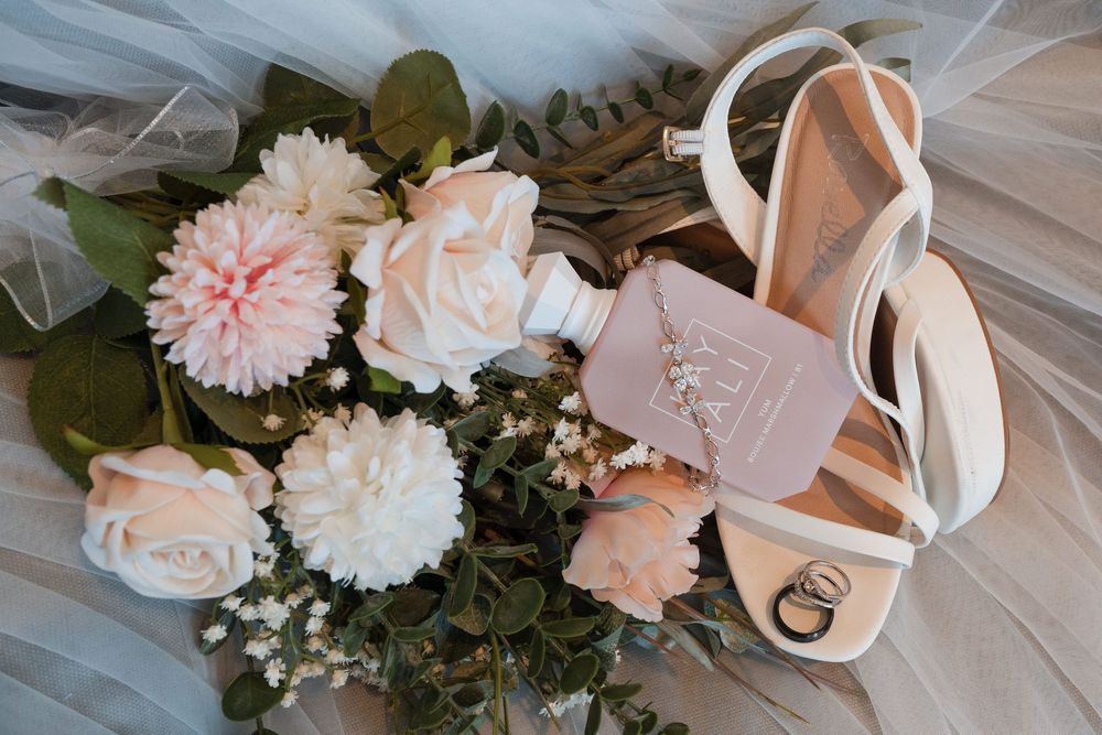 Bridal flat lay with blush roses, dahlias, white heels, wedding rings, and a pink perfume bottle on a veil.