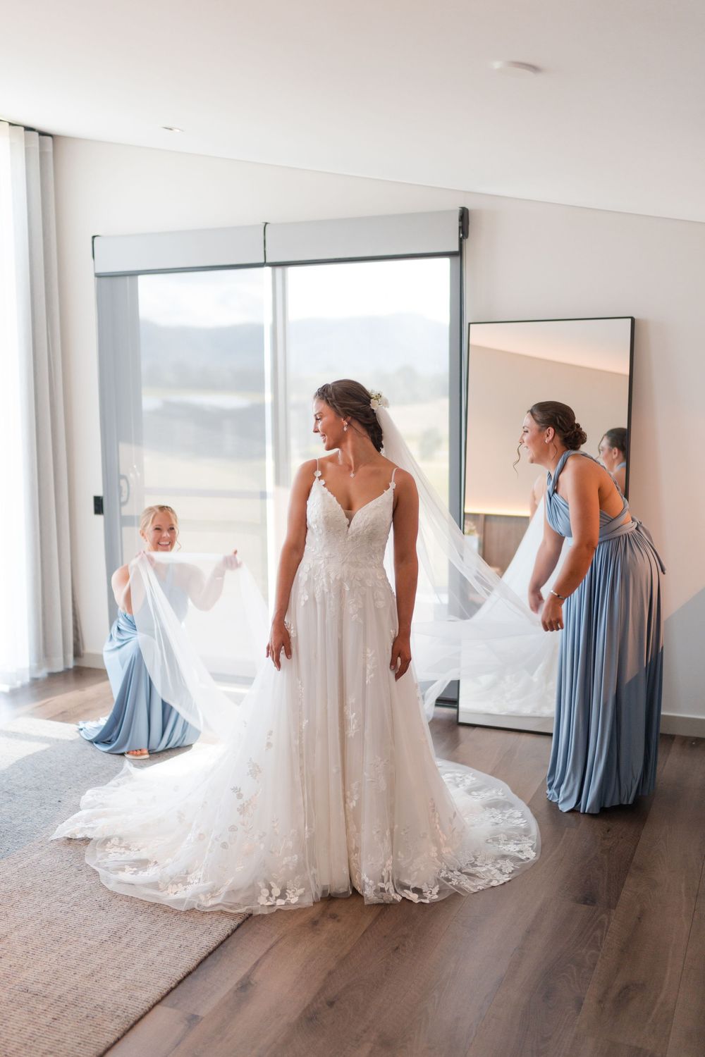 Bride in lace wedding gown with bridesmaids in blue dresses helping arrange veil in bright modern room.