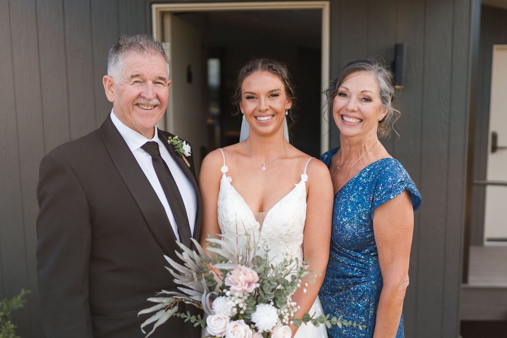 Bride in white lace gown holds bouquet flanked by smiling older couple in formal attire outside modern building.