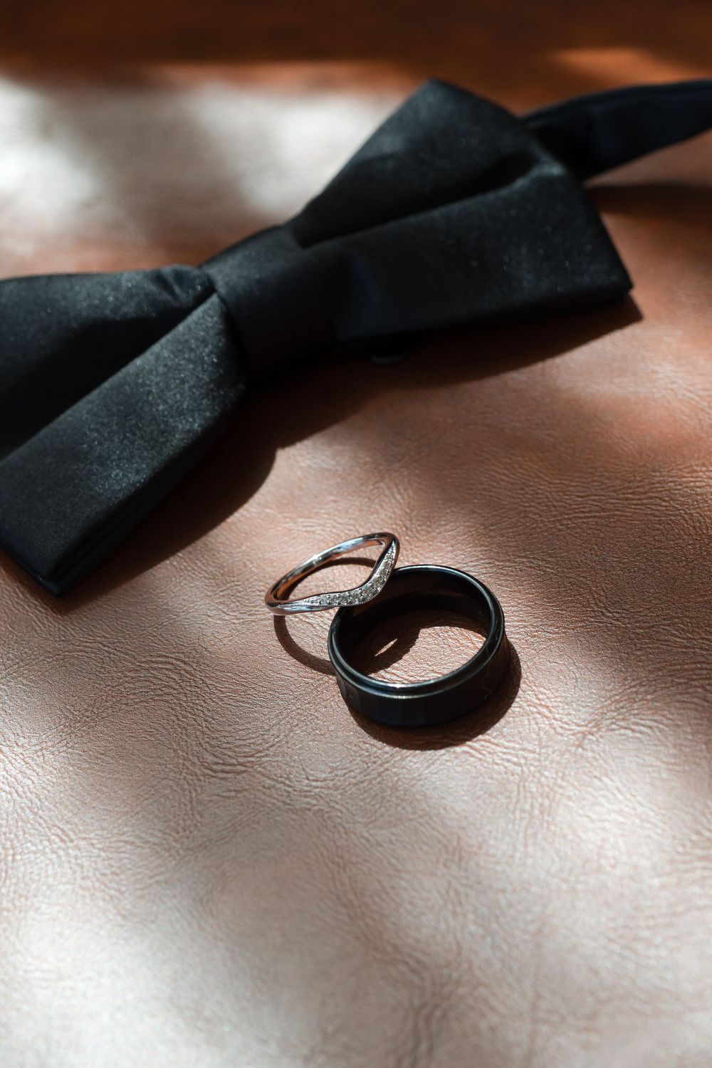 Two wedding rings beside a black satin bow tie on a beige leather surface in soft dramatic lighting.