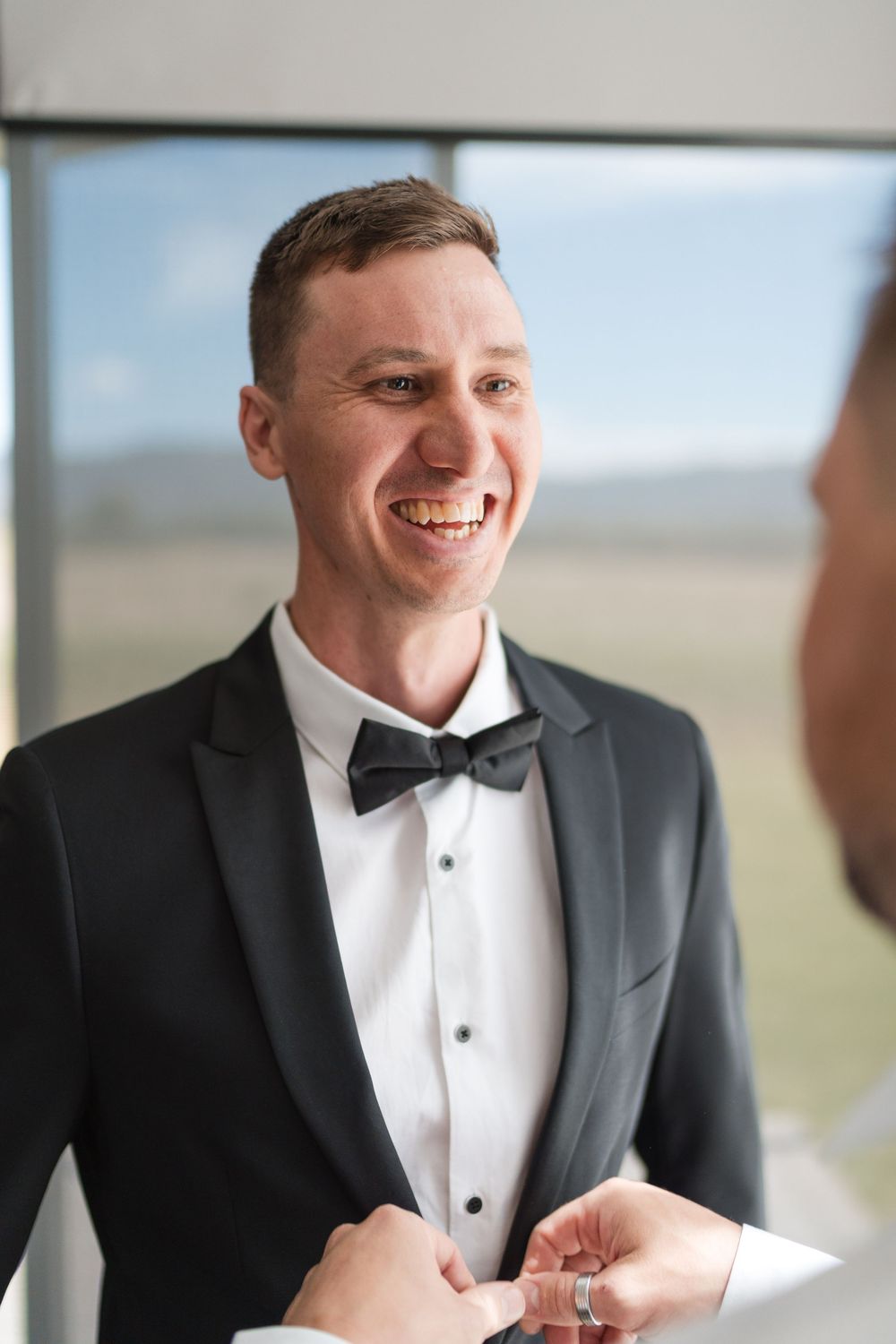 Smiling groom in black tuxedo and bow tie laughing with groomsman during wedding preparations indoors.