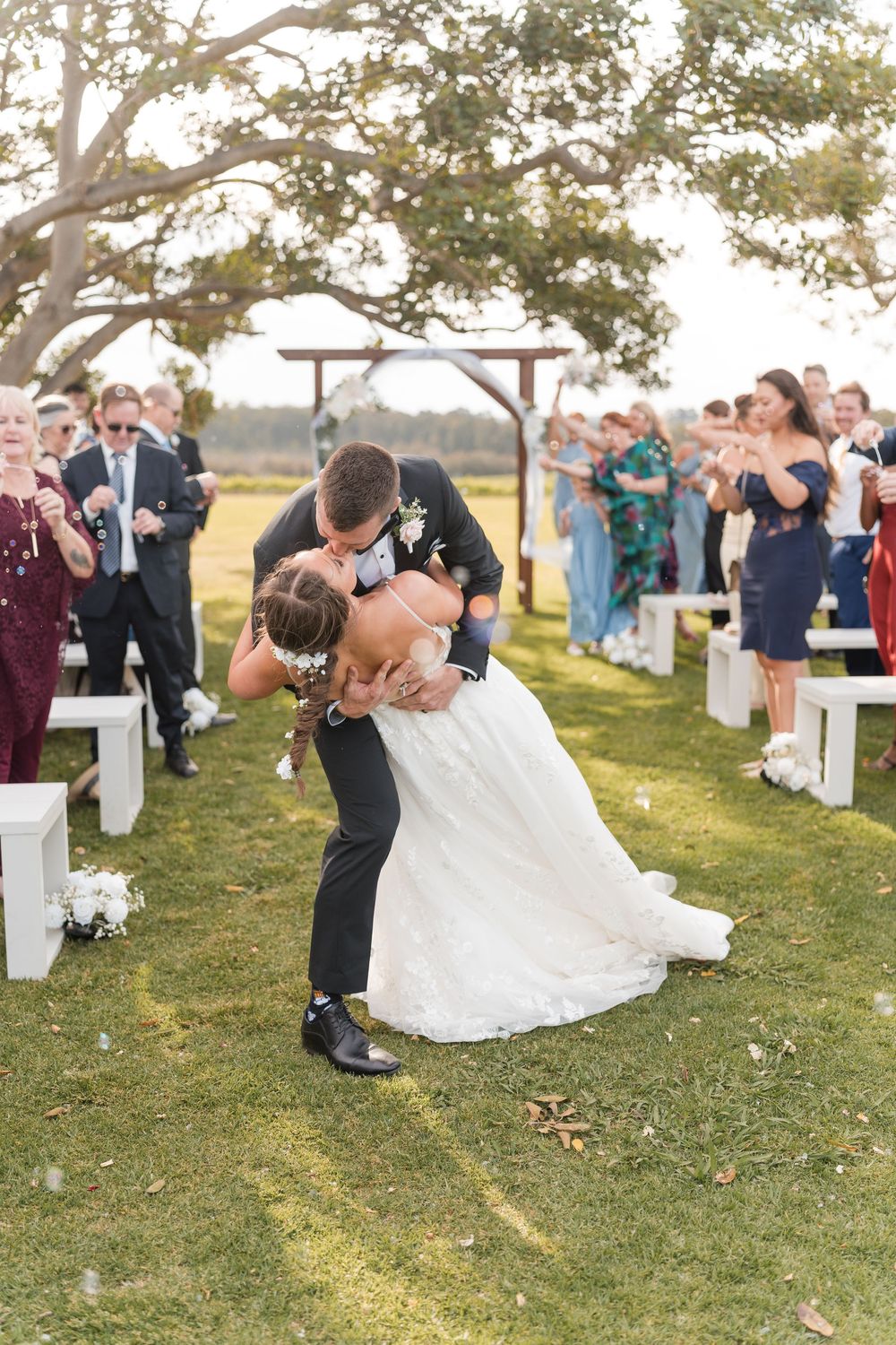 Groom dips bride for a kiss at outdoor wedding ceremony while guests celebrate along white bench aisle.