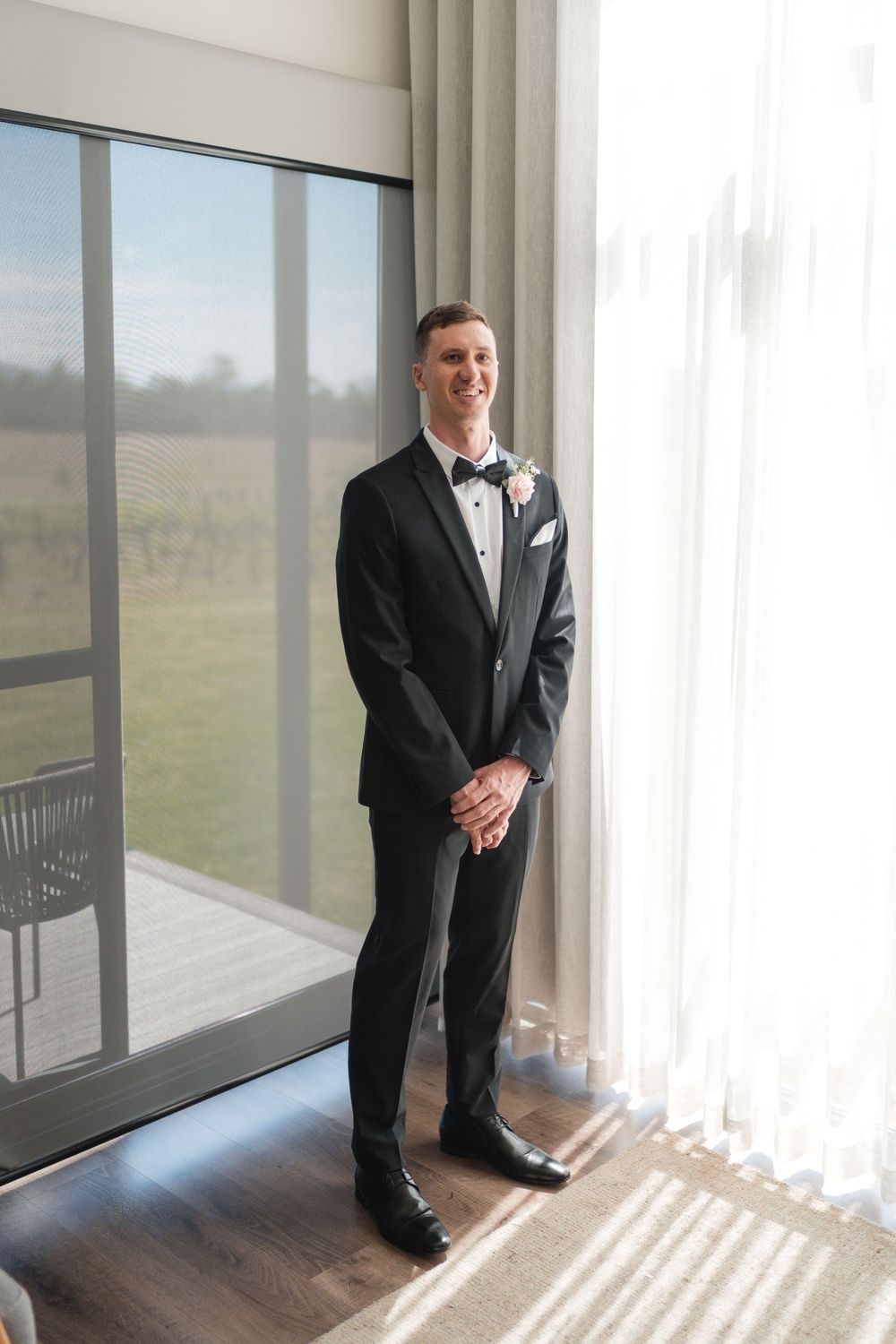 Smiling groom in black tuxedo with bow tie and boutonniere standing by large windows on his wedding day.