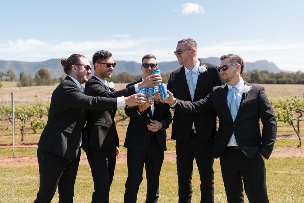Five groomsmen in black suits toasting with canned drinks in a sunny vineyard on a wedding day.