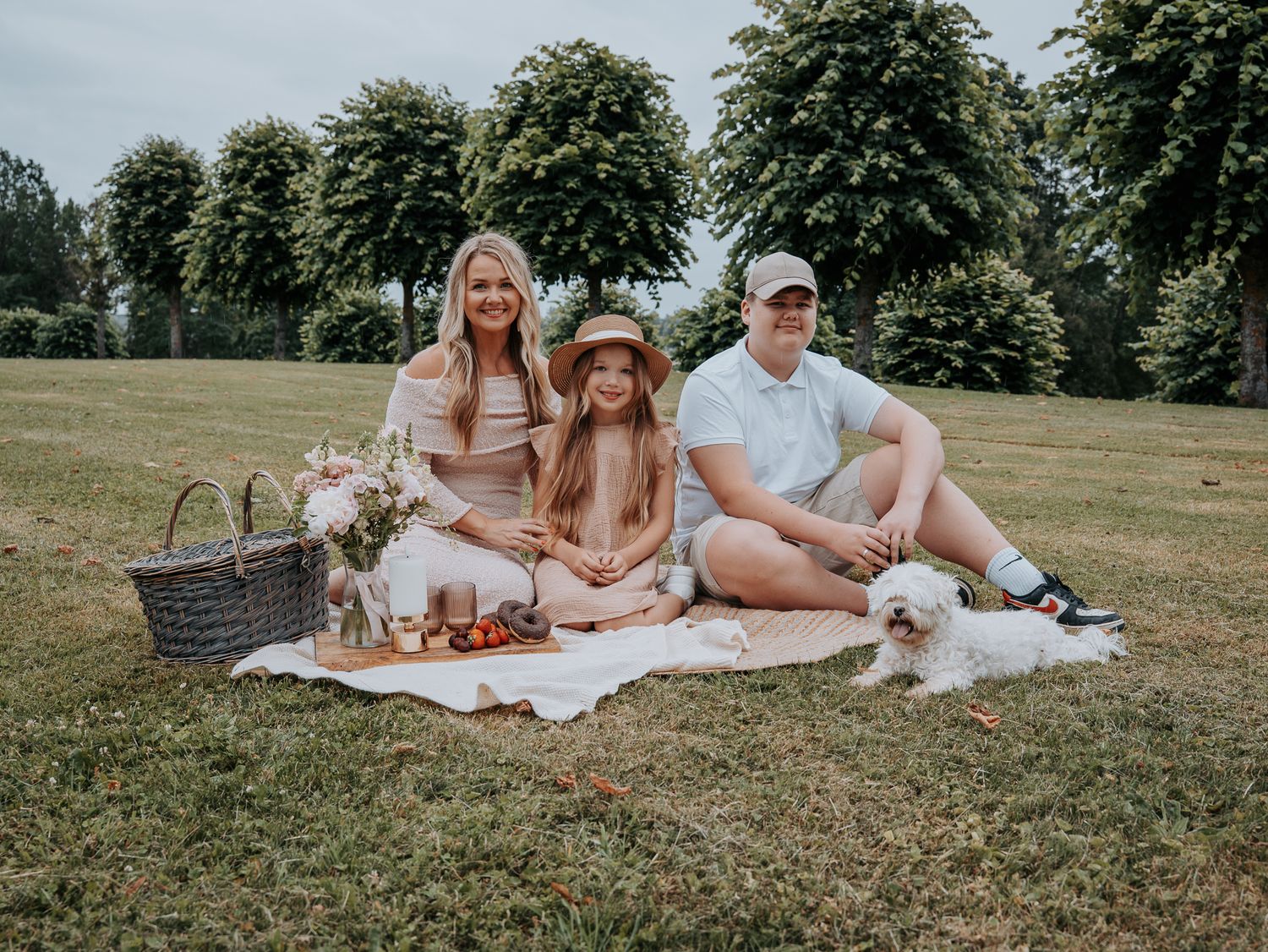 Family of three enjoying a picnic on a blanket in a lush green park with a small white dog.