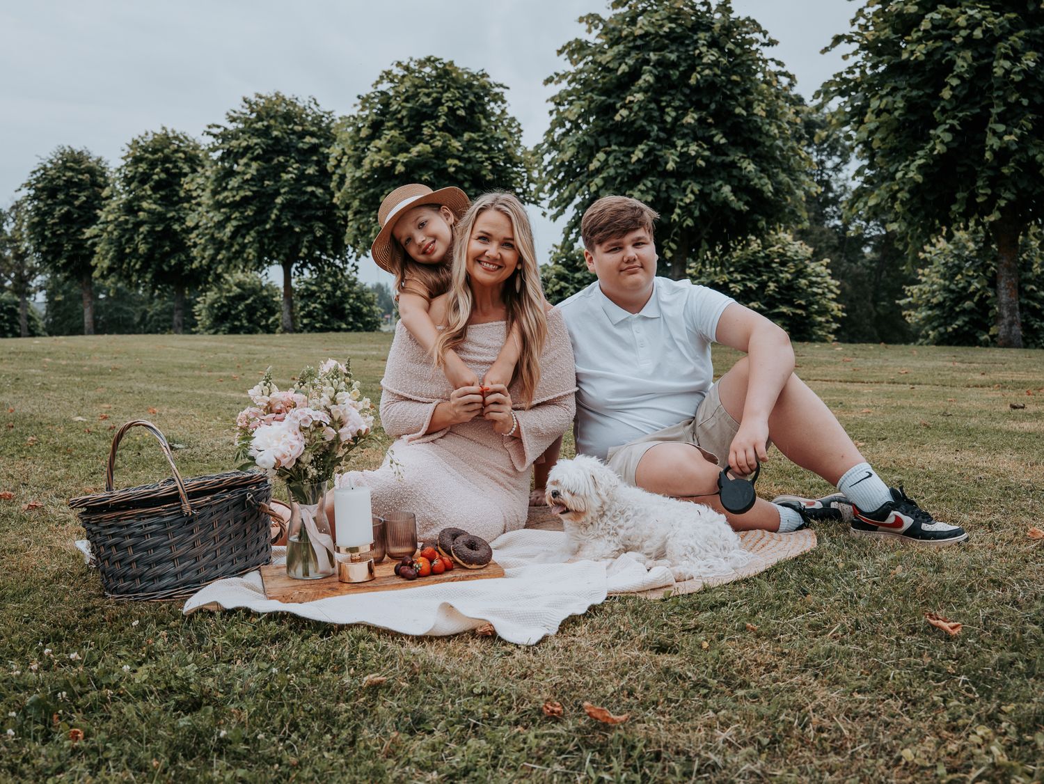 Three people enjoying a picnic on a blanket in a park with a small white dog, flowers, and a wicker basket.