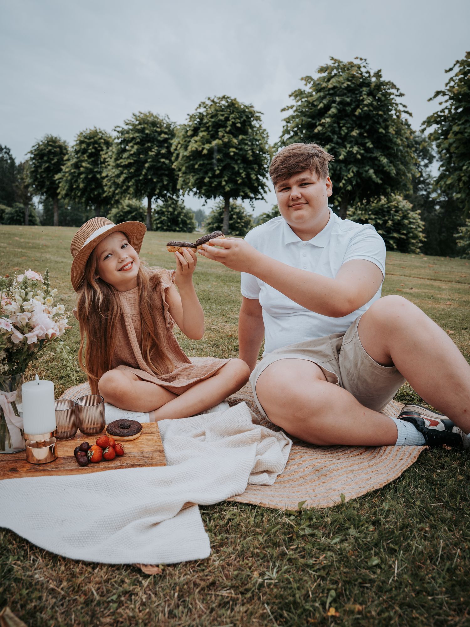 Two kids enjoying a picnic outdoors on a blanket in a lush green park, sharing food and smiling.