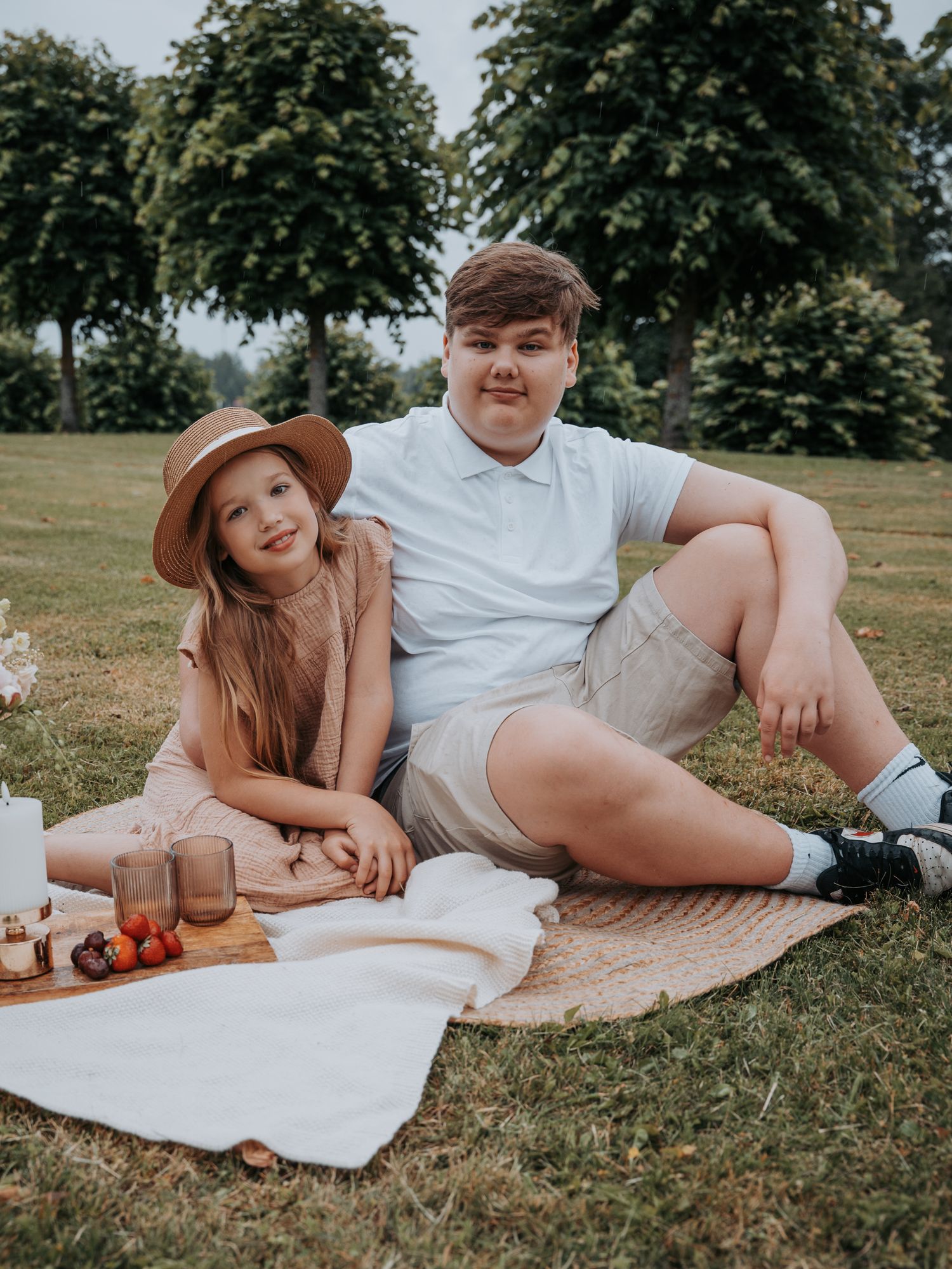 Two kids enjoying a sunny outdoor picnic on a blanket in a lush green park with food and drinks.