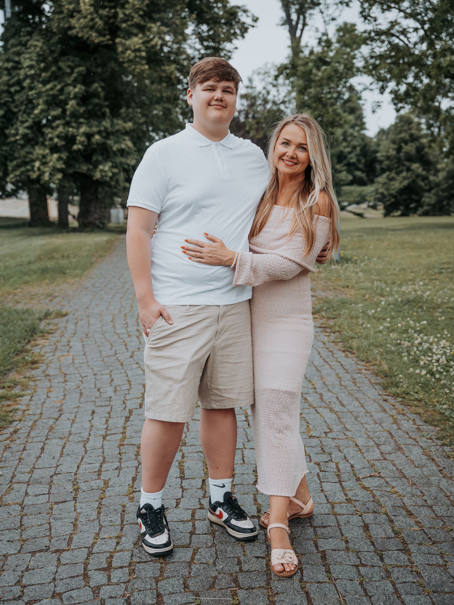 A young couple poses together on a brick pathway surrounded by lush green trees in a park setting.