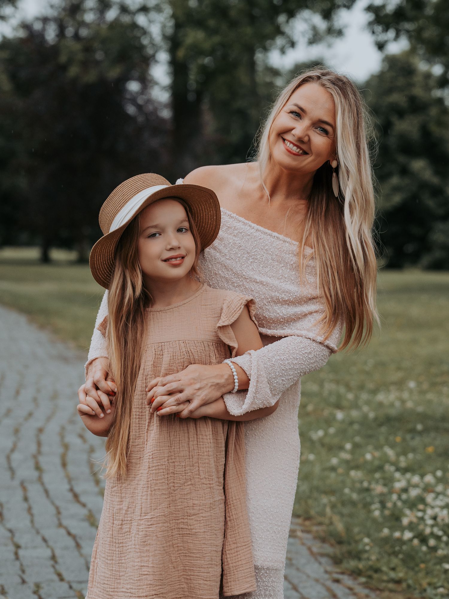 A smiling woman in an off-shoulder dress hugs a young girl in a hat and pink dress on a park path.