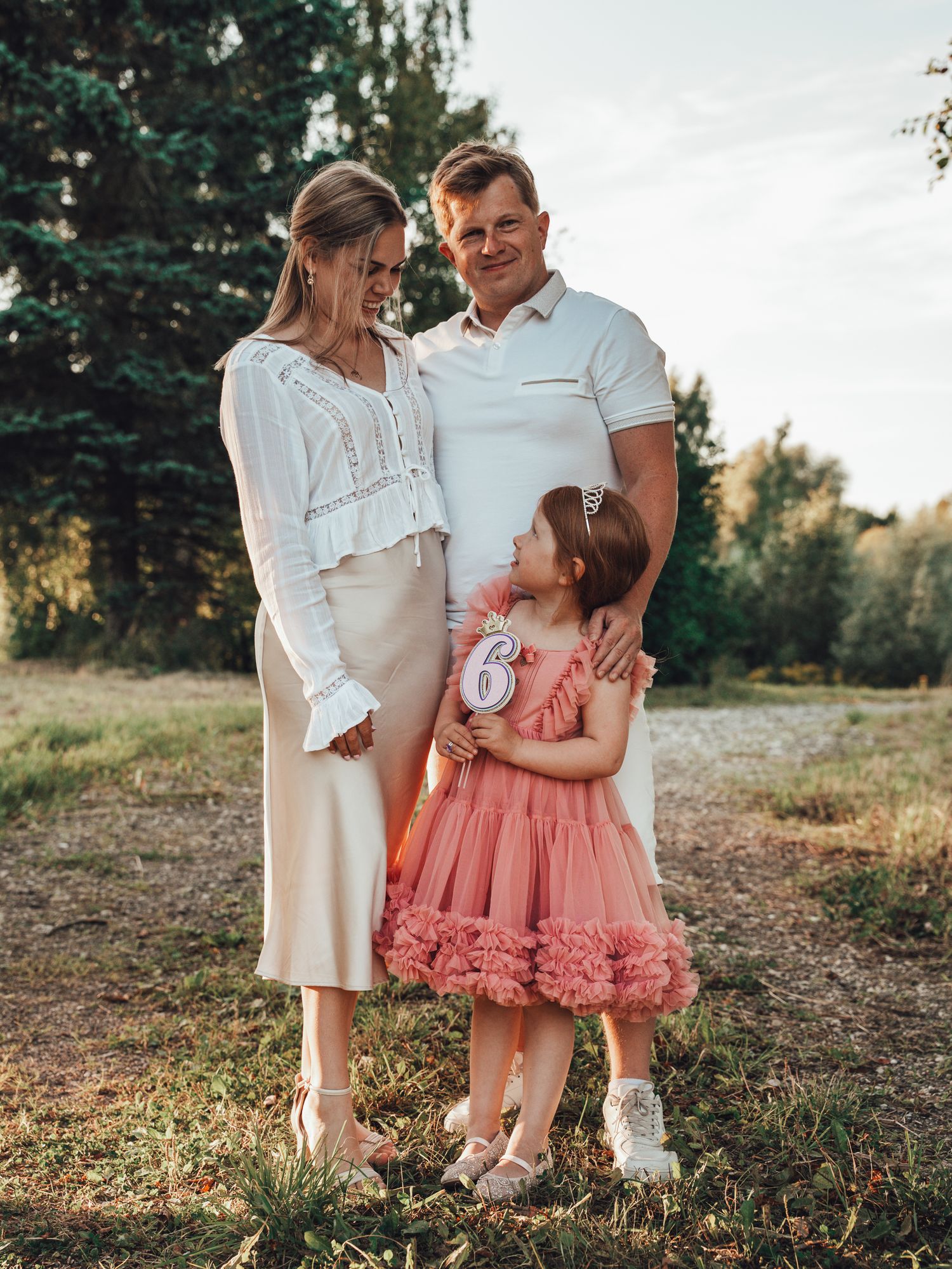 Family of three posing outdoors, parents in white outfits with young girl in pink tutu dress holding number 6 sign.