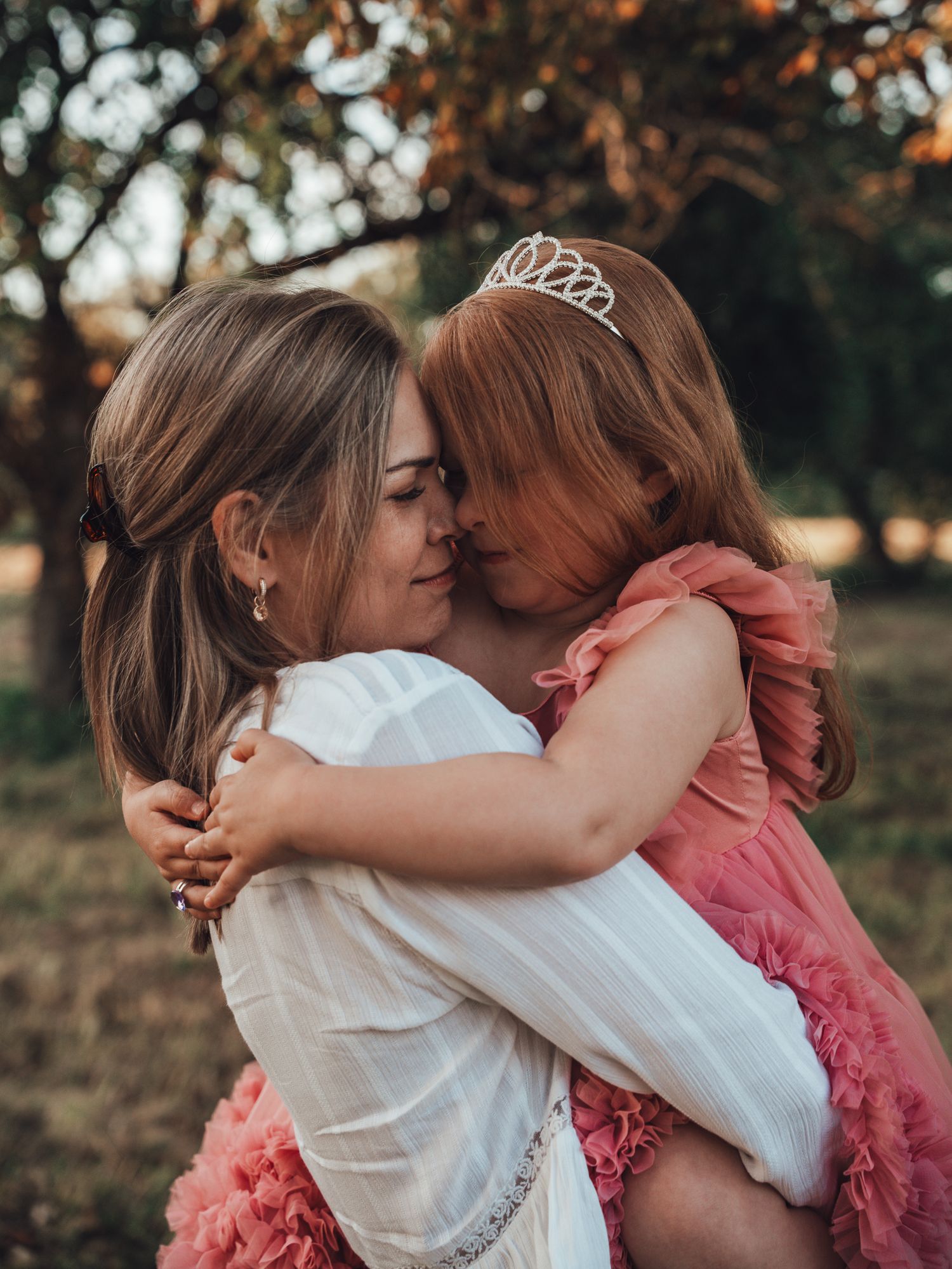 A woman in white dress holds a little girl wearing a pink tulle dress and tiara, sharing a tender embrace outdoors.