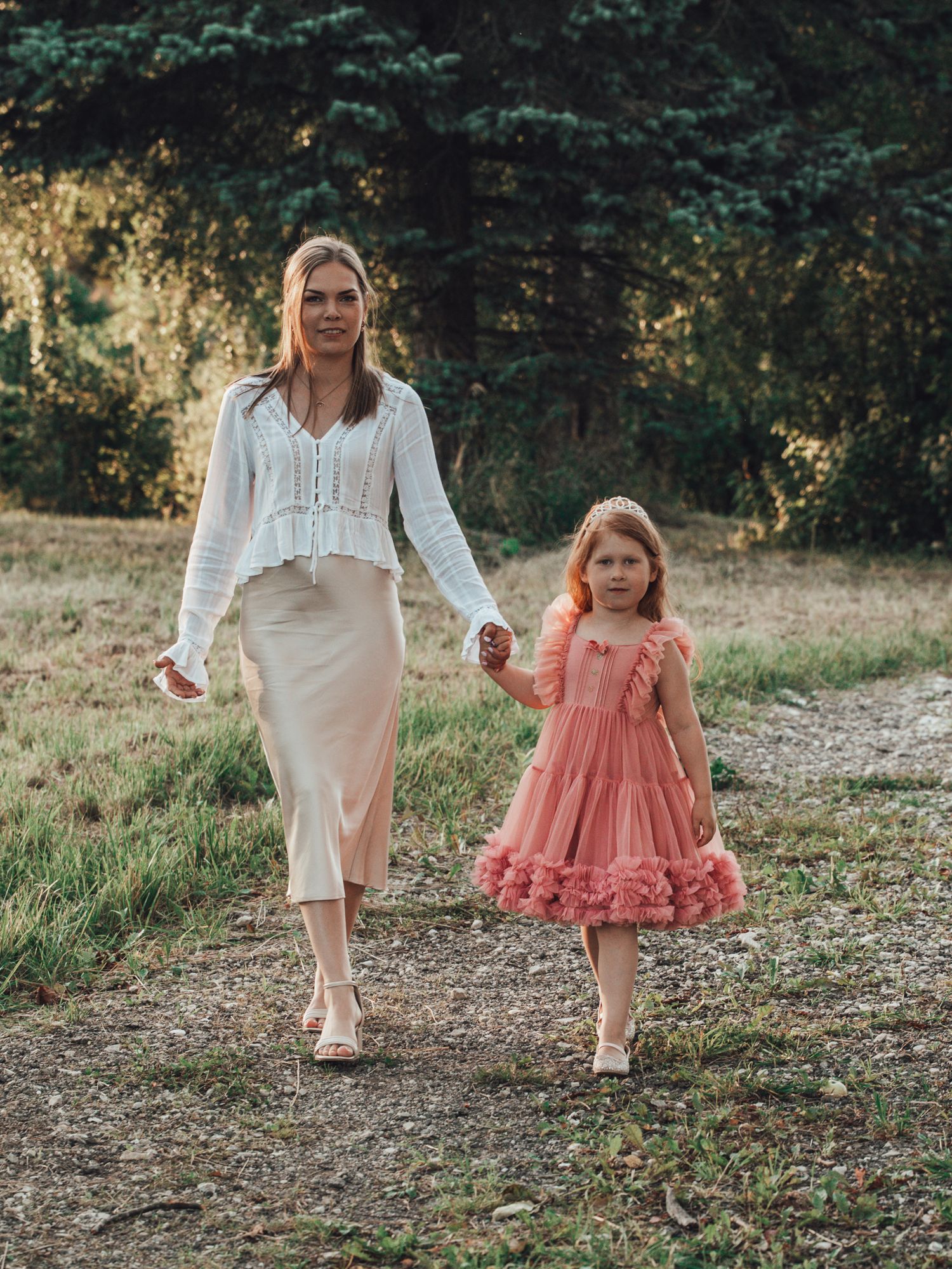A woman in a white blouse and beige skirt walks hand-in-hand with a young girl in a pink dress on a nature path.