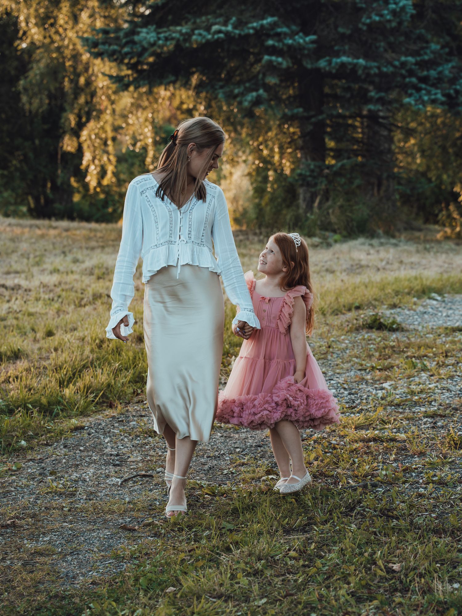 A woman in white and a little girl in pink dress walk hand-in-hand through a sunlit park path.