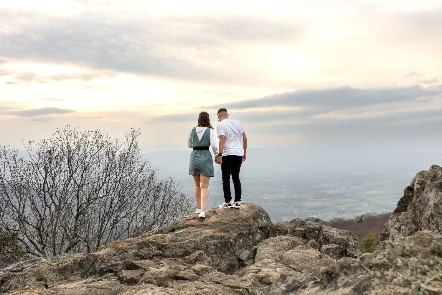 Two people stand together on a rocky cliff overlooking a misty mountainous landscape at sunset.