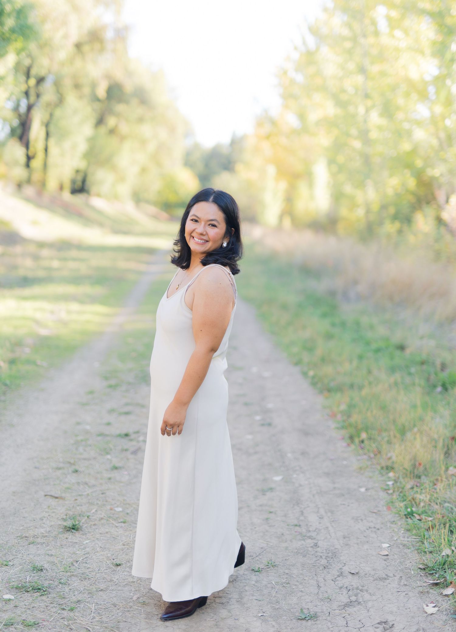 Woman in white maxi dress smiling on a sunlit dirt path surrounded by green trees in a fall mini session in North Davis with Jessica Heuer