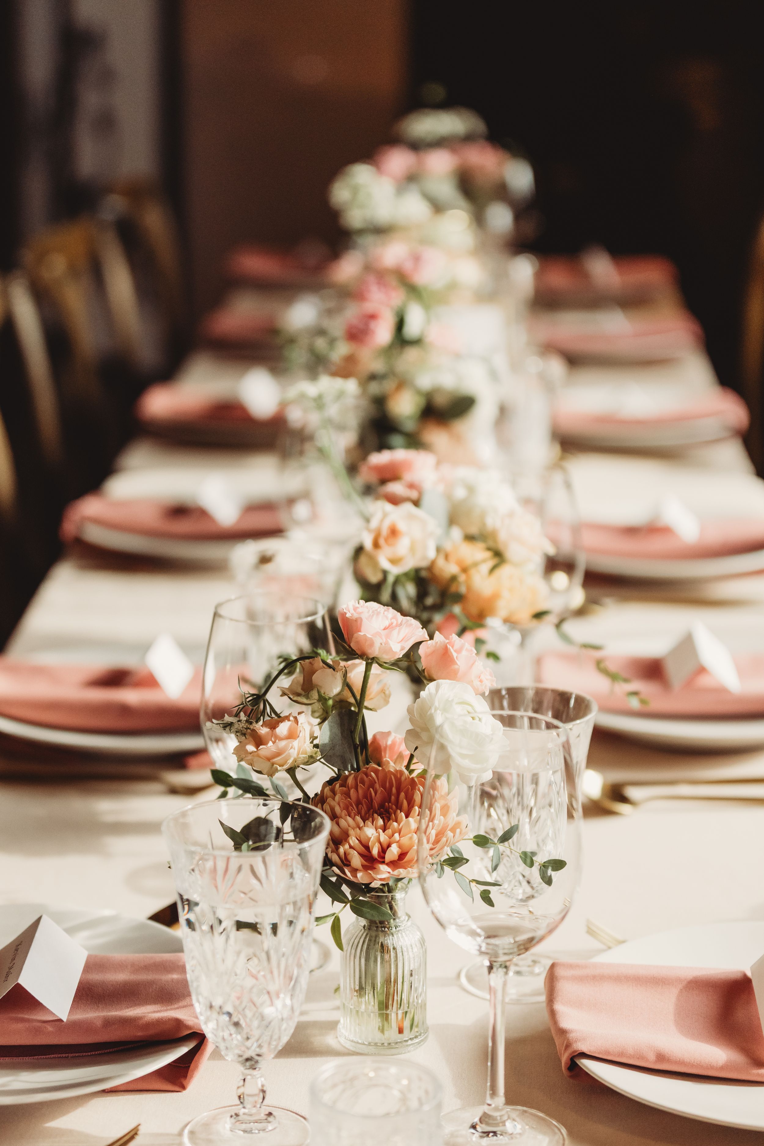 Elegant wedding reception table with pink napkins, crystal glasses, and floral centerpieces featuring roses and dahlias.