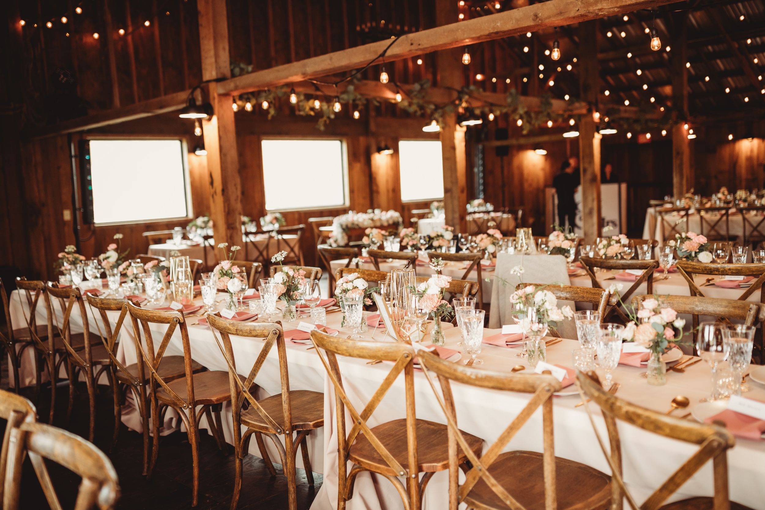 Rustic barn wedding reception with string lights, long tables, wooden cross-back chairs, and floral centerpieces.
