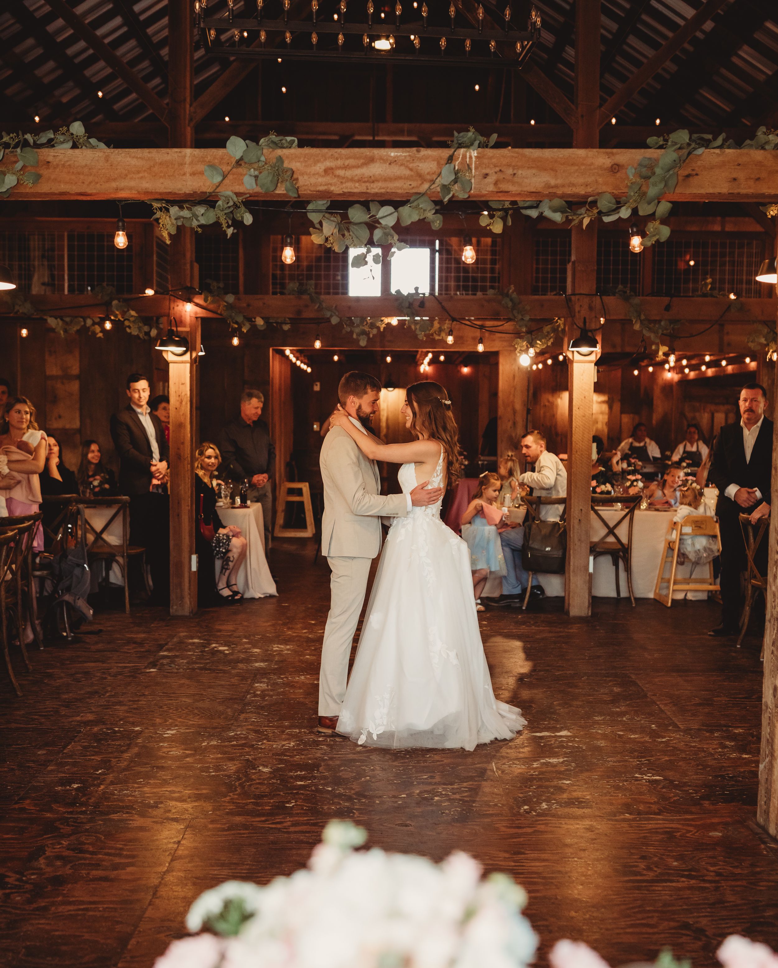 Bride and groom share first dance in rustic barn wedding venue with string lights, greenery garlands, and guests seated at tables.