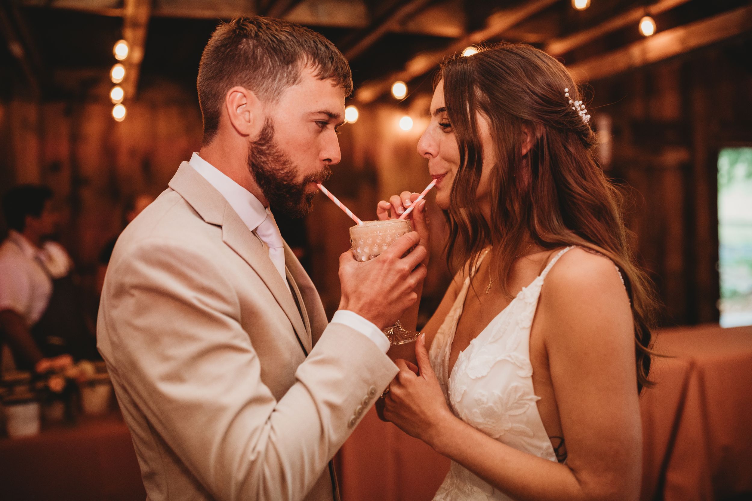 Bride and groom sharing a drink with straws at their rustic wedding reception with warm string lights.