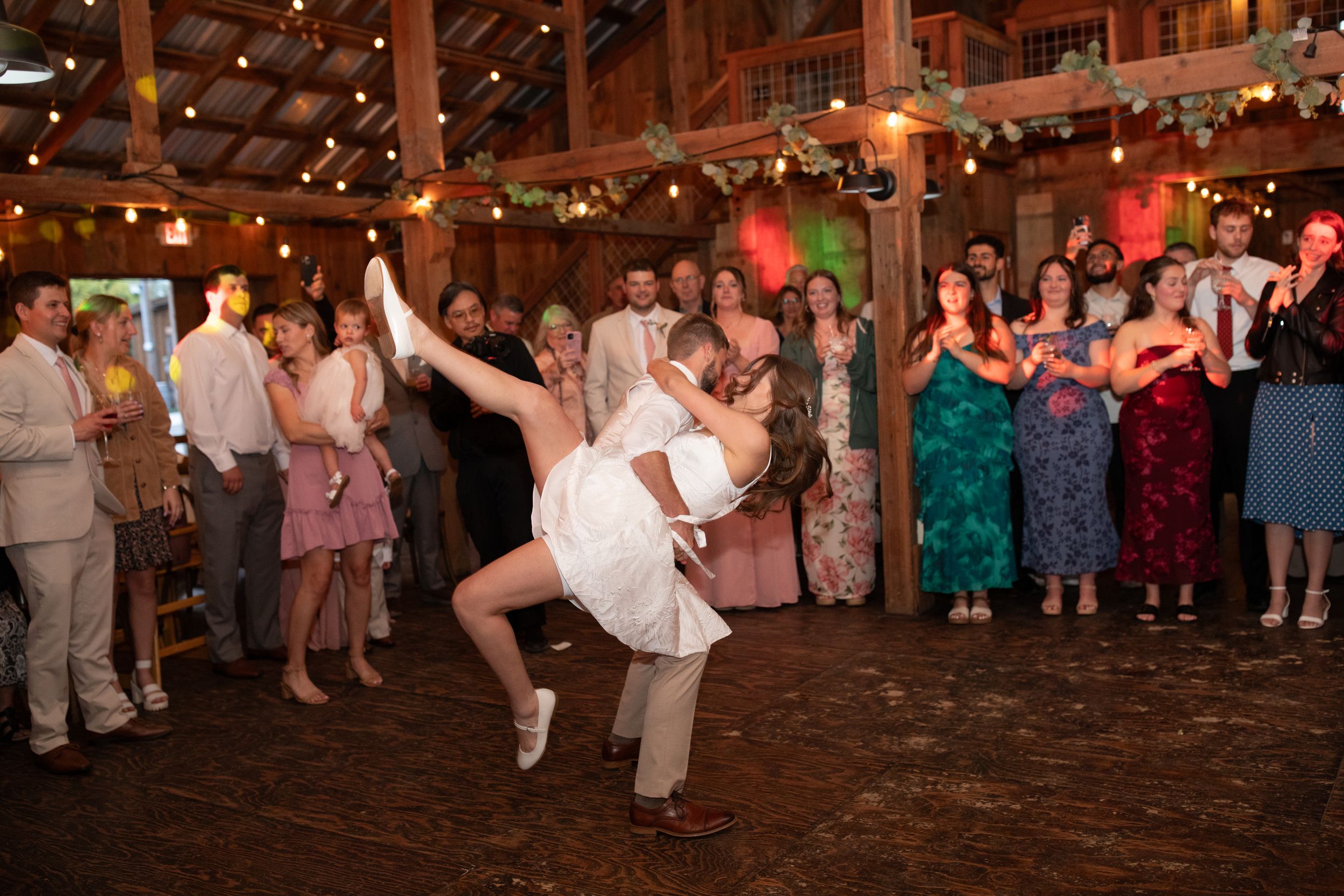 Bride and groom share a romantic dip dance at their rustic barn wedding reception with guests cheering.