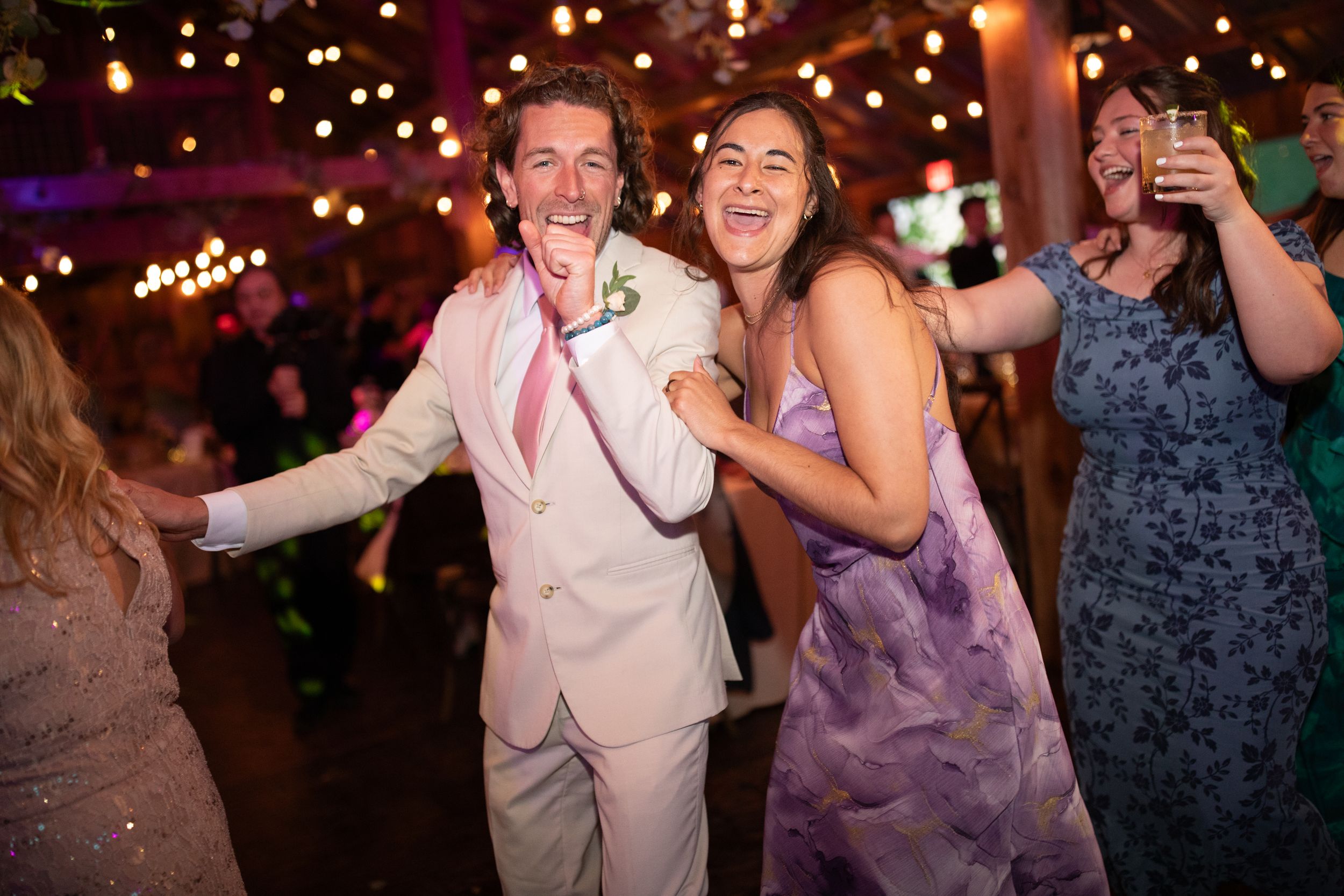 Couple dancing joyfully at a wedding reception with string lights and guests celebrating in the background.